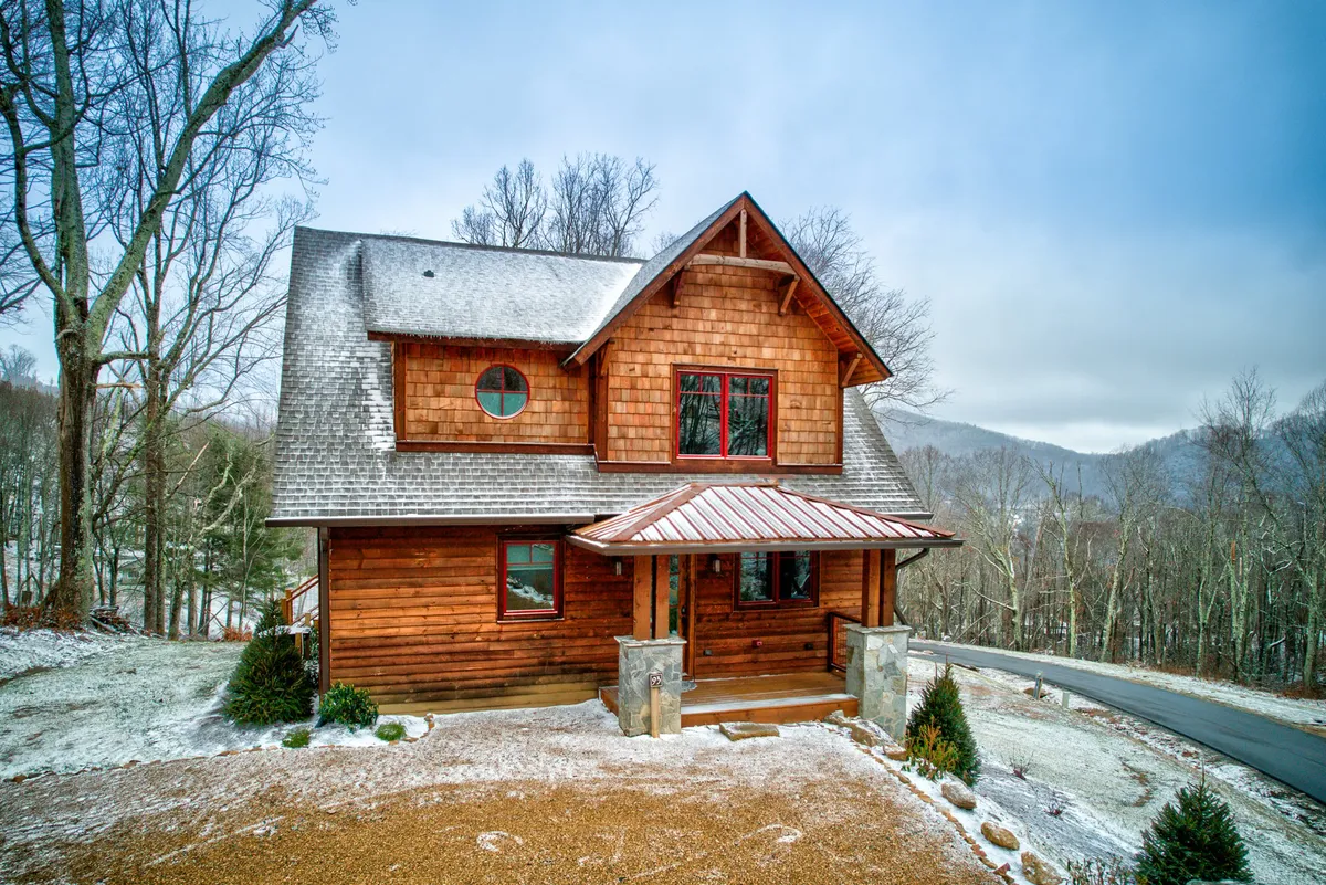 The image showcases the striking wooden exterior of Cornerstone Cabin, with expansive windows that invite natural light and offer glimpses of the surrounding forest and mountains. The architecture blends modern craftsmanship with rustic charm, set against a serene wooded backdrop—ideal for gathering outdoors or enjoying the view from the inviting porch.