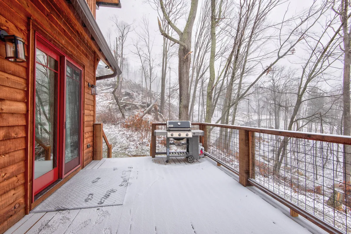A wooden deck equipped with a barbecue grill sits above a snowy ground, bordered by a line of trees. The grill and open design highlight the potential for outdoor cooking and dining, even in winter.
