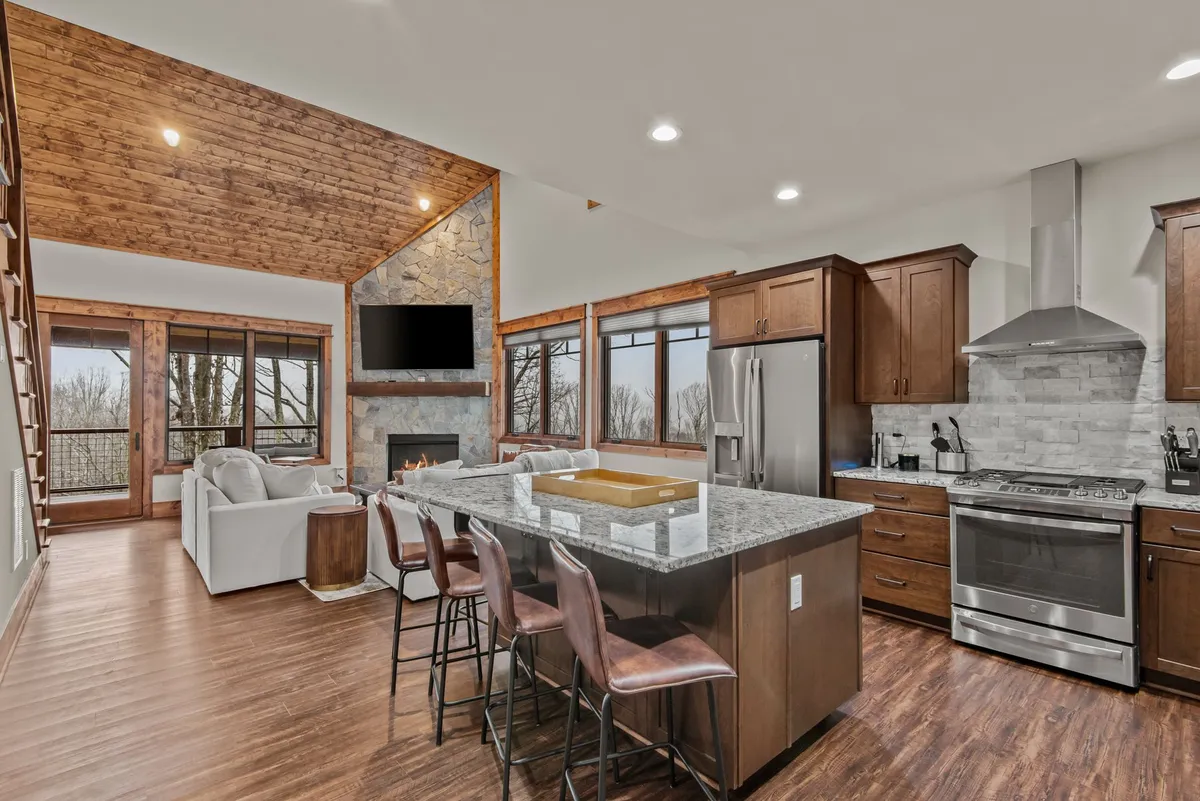 This image showcases an open-concept layout combining the kitchen and great room. It prominently features gleaming granite countertops, a stone fireplace, vaulted wooden beams, and expansive windows offering mountain and wooded views. The scene feels welcoming, highlighting opportunities for cooking, entertaining, and enjoying family gatherings.