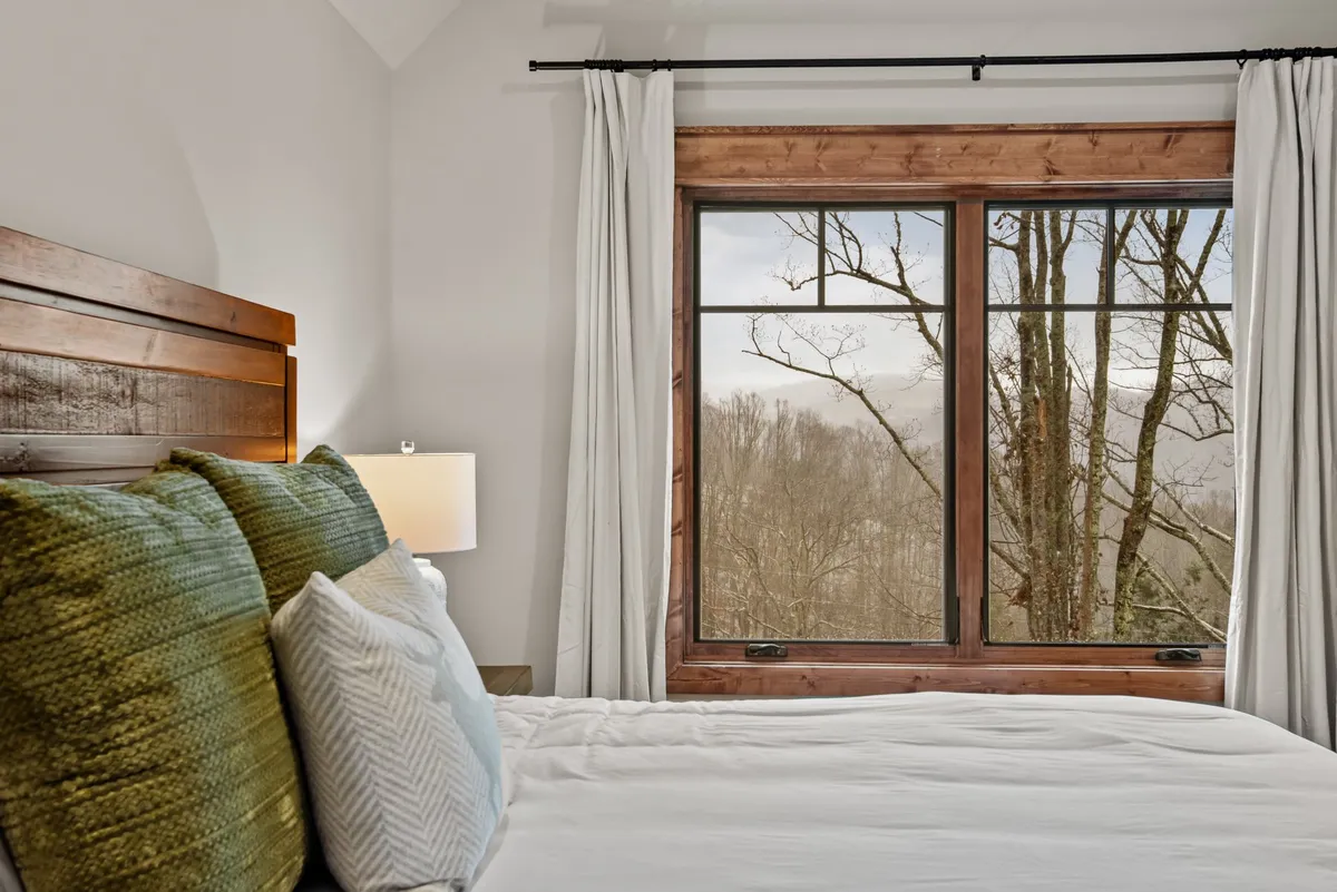A tranquil bedroom with a cozy bed outfitted in textured pillows and a large window offering a serene view of surrounding trees. The space is warm, comfortable, and ideal for unwinding after a day of adventure.
