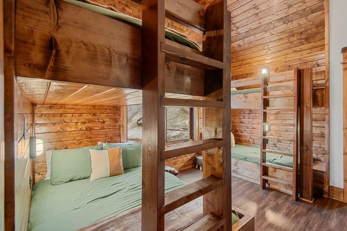 A cheerful bunk room with two sets of sturdy bunk beds, green bedding, a wooden ladder, and ample natural light streaming through large windows. The playful space is designed for kids or groups, offering both comfort and togetherness.