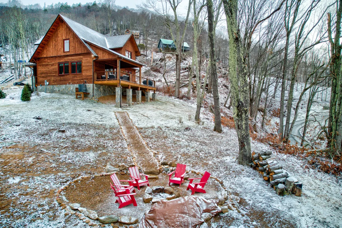Red Adirondack chairs circle a stone fire pit outside a wooden cabin, set among tall trees. The scene captures a communal mood, inviting guests to gather, share stories, and enjoy the mountain night air around the fire.