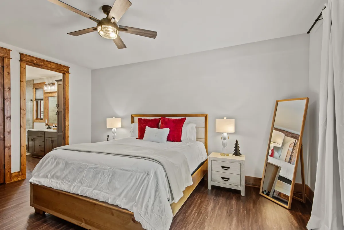 A spacious bedroom featuring a plush bed adorned with red and white pillows, flanked by wooden nightstands with lamps. The room has a full-length mirror, a wooden ceiling fan, and rustic wooden accents, creating a serene and elegant atmosphere ideal for rest and relaxation.