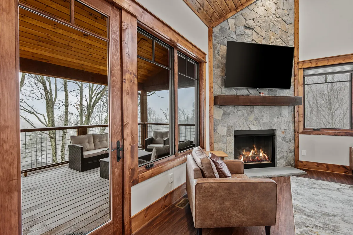 A spacious living area with a stone fireplace, large windows overlooking the deck, comfortable seating, and a TV mounted on the wall. The visible deck suggests easy indoor-outdoor flow, ideal for group activities.