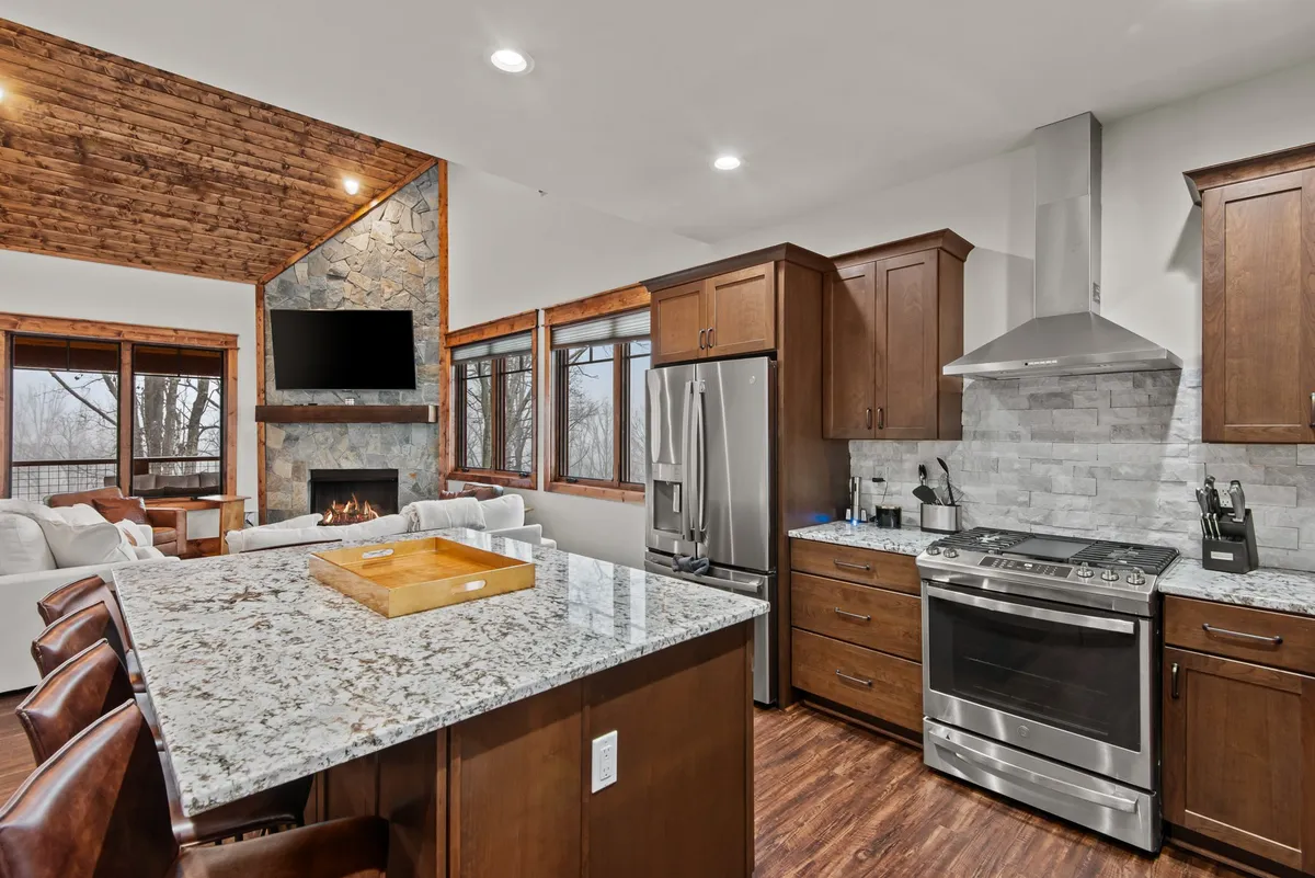The photo highlights a spacious kitchen area with granite countertops, stainless steel appliances, a stone fireplace, and generously sized windows letting in natural light. The space feels both functional and inviting, perfect for group cooking, entertaining, and relaxing by the fire with beautiful views in sight.
