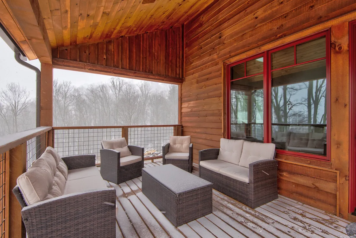 This image captures an inviting outdoor seating area on a wooden deck, with comfortable chairs, a wooden railing, and large windows that look back into the home. The surrounding landscape is tranquil and the atmosphere is peaceful, perfect for relaxing outdoors or enjoying the mountain air.