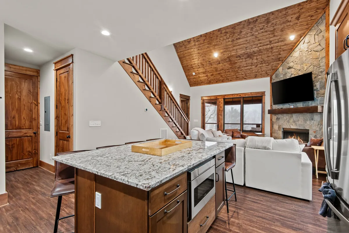 A warm, inviting interior with a central granite kitchen island, stone fireplace, soaring wooden ceiling, and large windows framing natural mountain views. The space is set for both cooking and relaxing, with a cozy ambiance ideal for gatherings after a day’s adventure.