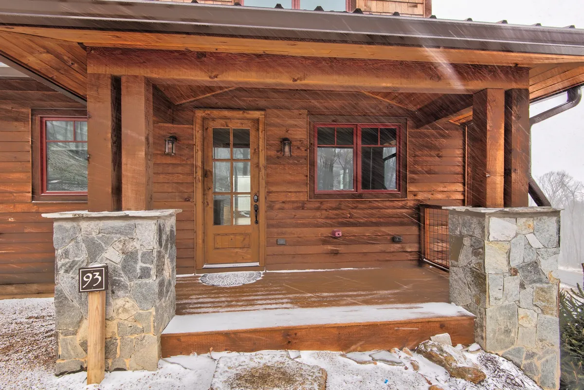 This image showcases the exterior entrance of the cabin, featuring a welcoming wooden door, stone pillars, and distinctive red window frames. The scene reflects a sense of arrival and the potential for gathering with friends, especially during festive or seasonal occasions.