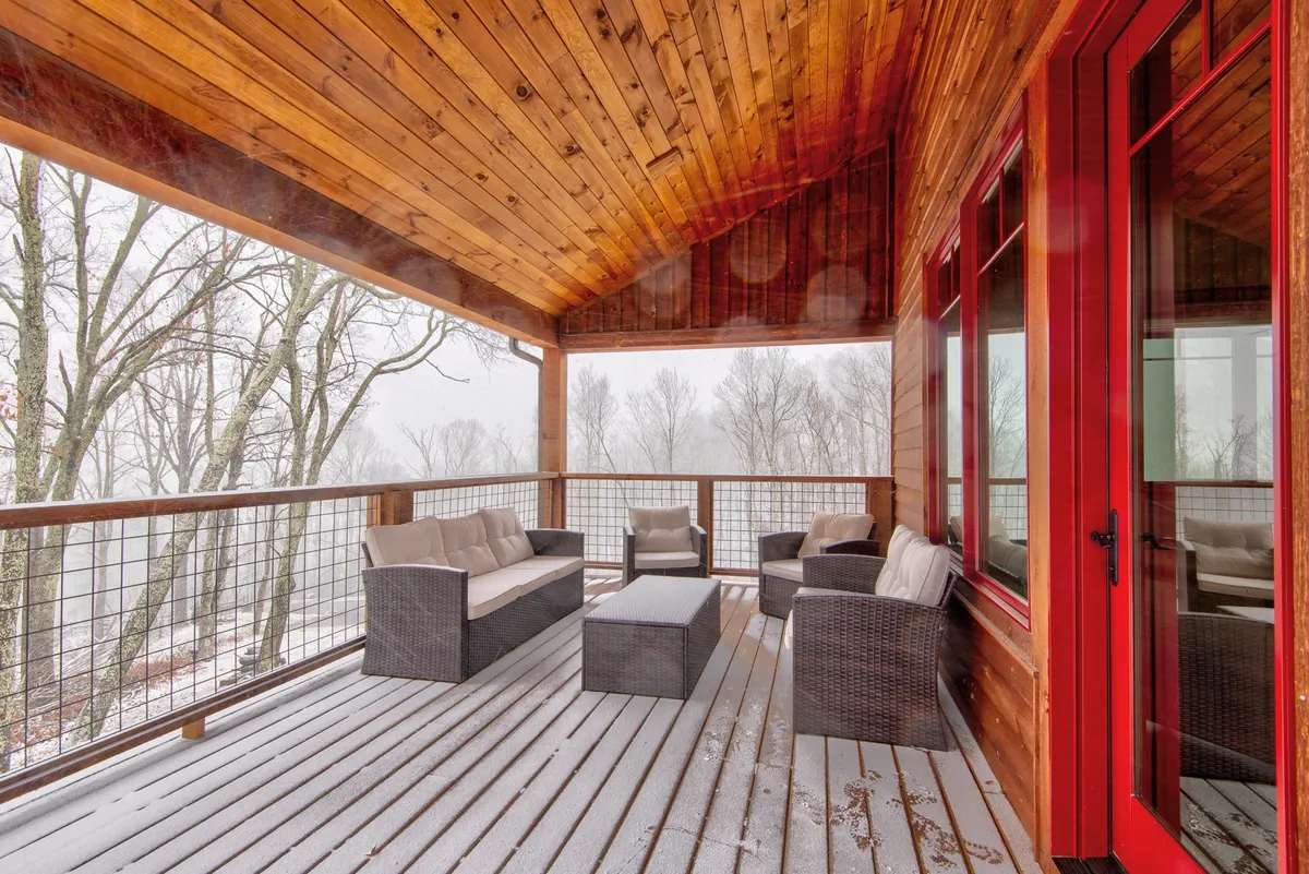 A covered outdoor seating area with a wooden ceiling, red-framed windows, and comfortable outdoor furniture. The space invites guests to relax outside while remaining sheltered, even as snow falls outside.