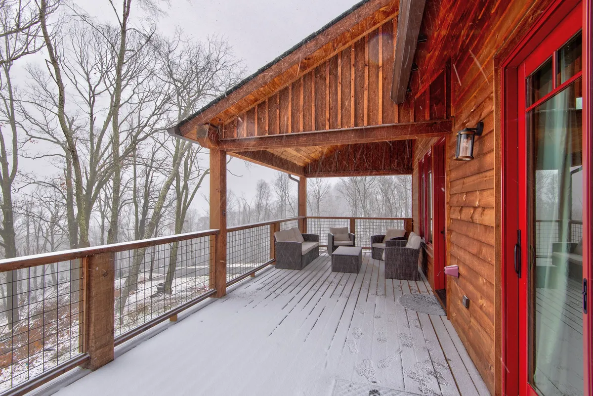 The image shows a spacious wooden deck surrounded by snow, with cushioned seating arranged for comfort. The deck overlooks a tranquil winter landscape, offering an inviting area for relaxing with a hot drink or gathering with loved ones.